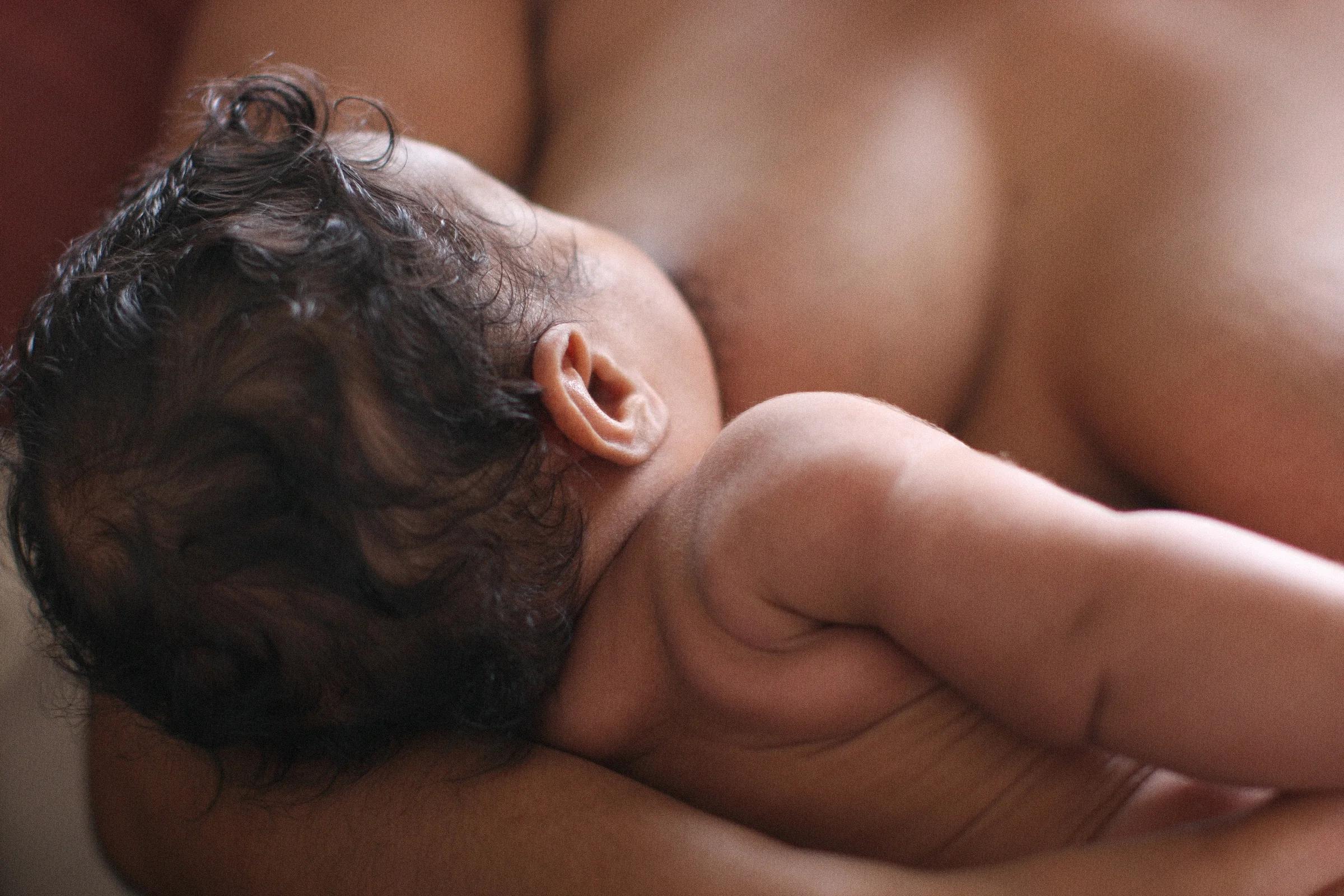 A baby feeding at the breast, cradled in a parent’s arm, in warm window light